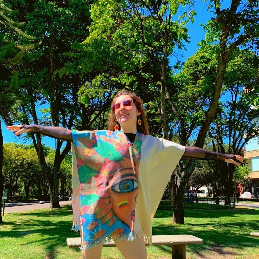 Mujer con los brazos abiertos luciendo un poncho artesanal ilustrado, en un parque