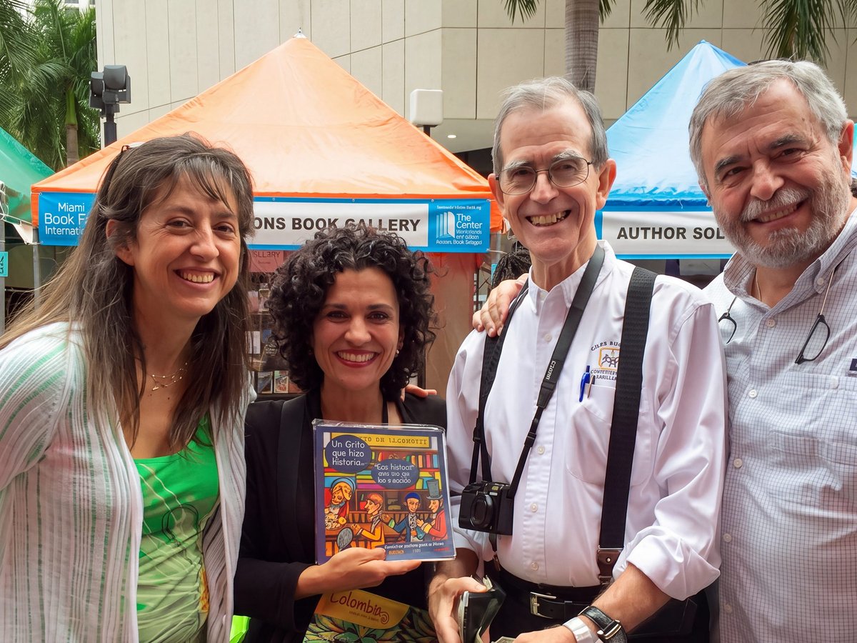 Cuatro personas sonríen frente a los stands de la Feria del Libro de Miami; una sostiene la cartilla ilustrada «Un grito que hizo historia»